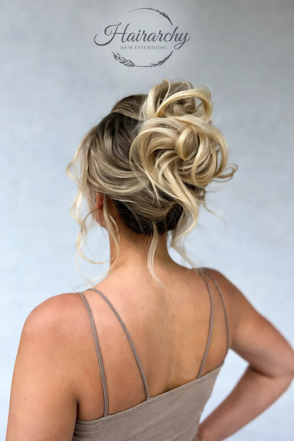 Back view of a woman with a voluminous blonde messy bun hairstyle with soft curled strands and blended highlights, styled with hair extensions, on a light neutral background with Hairarchy Hair Extensions logo at the top.