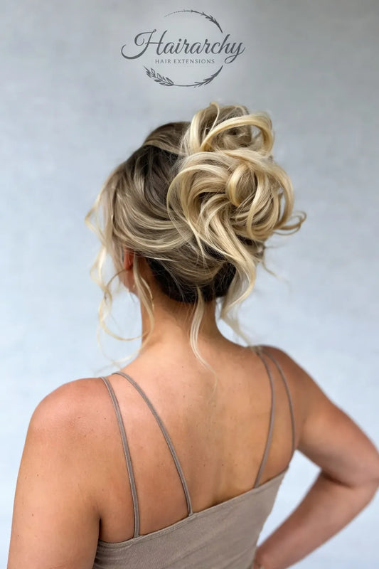 Back view of a woman with a voluminous blonde messy bun hairstyle with soft curled strands and blended highlights, styled with hair extensions, on a light neutral background with Hairarchy Hair Extensions logo at the top.